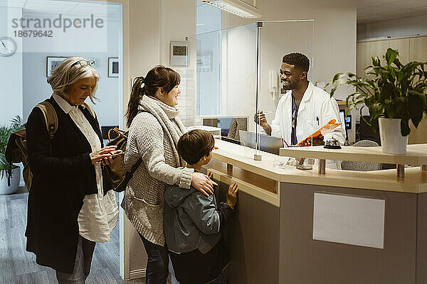 Stehende Patienten  die sich durch ein transparentes Schild mit einem männlichen Rezeptionisten in einer Klinik unterhalten