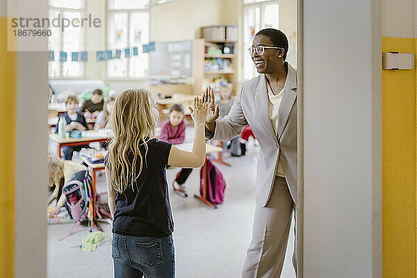 Lächelnde Lehrerin und Schulmädchen geben sich an der Tür im Klassenzimmer die Hand