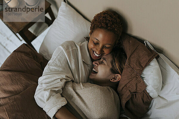 Glückliches nicht-binäres Paar  das zusammen lacht  während es zu Hause auf dem Bett liegt