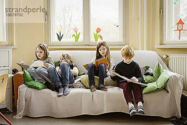 Mädchen und Junge in voller Länge beim Lesen von Büchern  während sie auf einem Sofa im Klassenzimmer sitzen