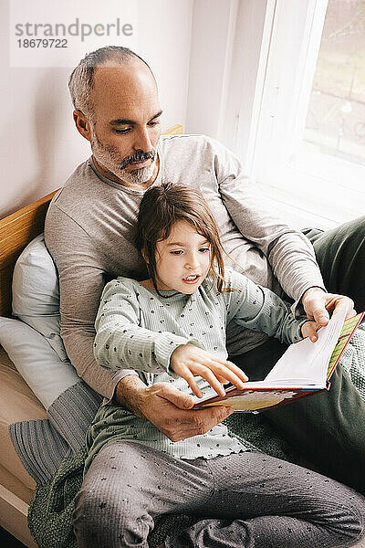 Vater und Tochter lesen ein Buch  während sie zusammen auf einer Fensternische zu Hause sitzen