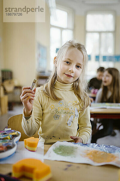Porträt einer blonden Schülerin mit Pinsel im Klassenzimmer