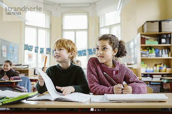 Lächelnder blonder Junge  der mit einem Mädchen am Schreibtisch sitzt und im Klassenzimmer wegschaut