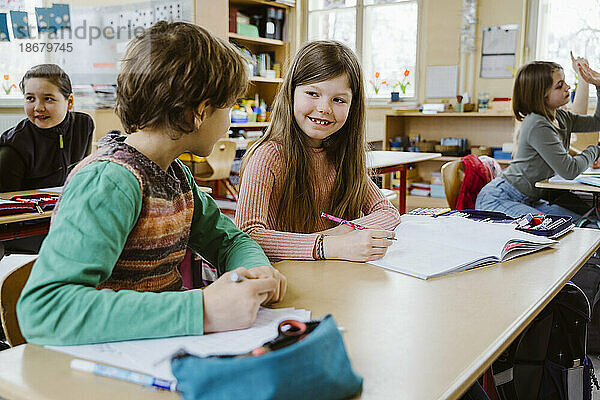 Lächelndes Mädchen im Gespräch mit einem männlichen Freund  während sie am Schreibtisch im Klassenzimmer sitzt