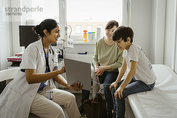 Eine Kinderärztin zeigt einer Mutter und ihrem Sohn  die im Untersuchungsraum einer Klinik sitzen  einen Tablet-PC