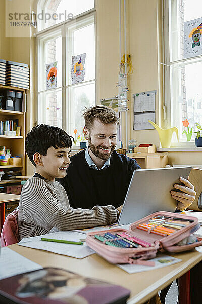 Zufriedener männlicher Lehrer  der einem Schüler bei der Benutzung eines Tablet-PCs am Schreibtisch in der Schule hilft