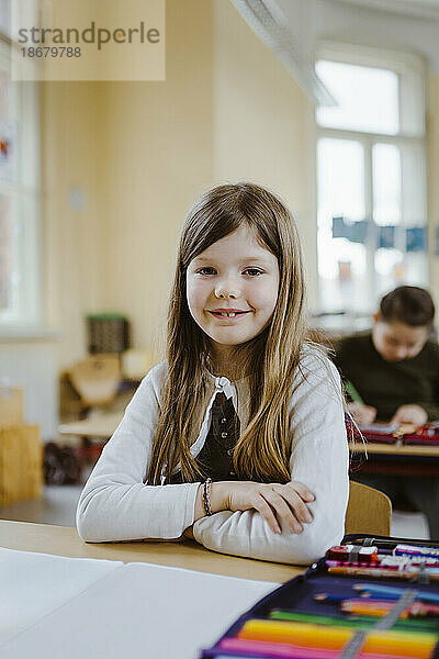 Porträt einer lächelnden Schülerin  die mit verschränkten Armen am Schreibtisch im Klassenzimmer sitzt