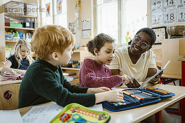 Lächelnder Lehrer  der einem Jungen und einem Mädchen mit einem digitalen Tablet hilft  die am Schreibtisch im Klassenzimmer sitzen