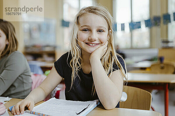 Lächelndes blondes Schulmädchen  das sich auf den Ellbogen stützt  während es am Schreibtisch im Klassenzimmer sitzt