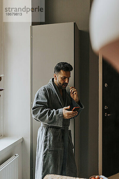 Mann im Bademantel benutzt Smartphone beim Zähneputzen zu Hause