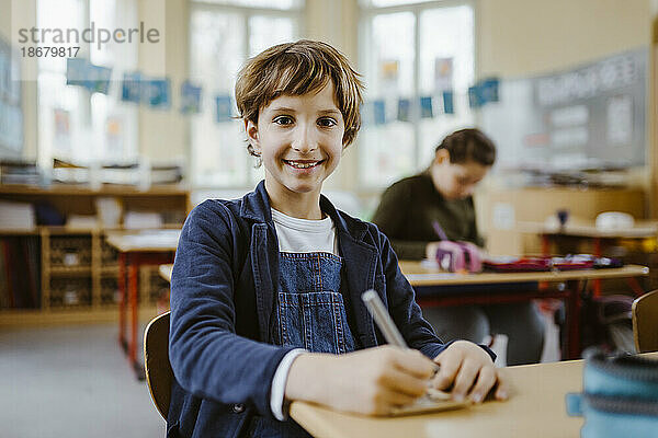 Porträt eines lächelnden Schülers  der im Klassenzimmer am Schreibtisch sitzt