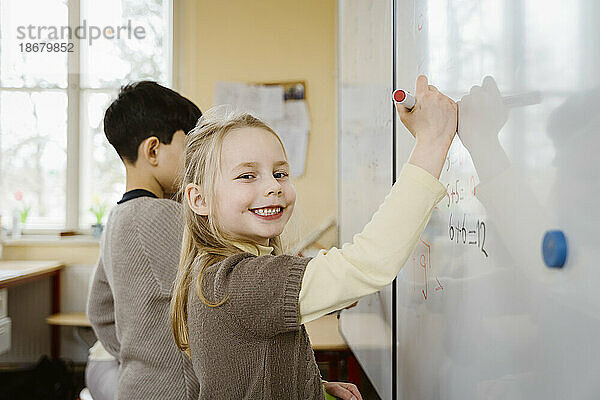 Porträt eines lächelnden blonden Mädchens  das auf einer Tafel im Klassenzimmer schreibt