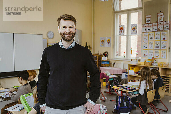 Porträt eines lächelnden bärtigen Lehrers  der vor Schülern im Klassenzimmer steht