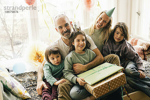 Porträt eines glücklichen Geburtstagskindes  das mit Geschenken und Familie auf einem Nischensitz zu Hause sitzt