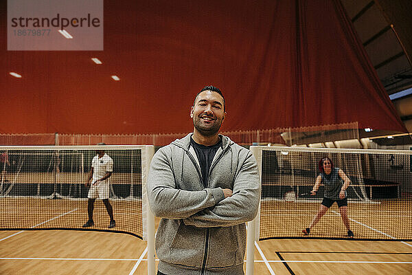 Lächelnder männlicher Trainer  der mit verschränkten Armen auf dem Badmintonplatz steht