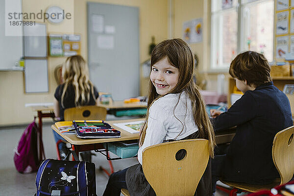 Porträt eines lächelnden Mädchens  das über die Schulter schaut und auf einem Stuhl neben einem Freund im Klassenzimmer sitzt
