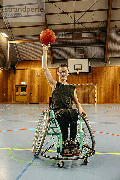 Lächelndes Mädchen  das im Rollstuhl sitzend auf dem Sportplatz einen Basketball balanciert