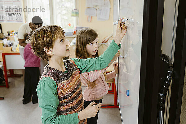 Junge schreibt auf Tafel  während ein Mädchen im Klassenzimmer eine Matheaufgabe löst