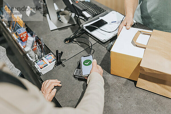Ausgeschnittenes Bild eines männlichen Kunden  der ein Kreditkartenlesegerät über sein Smartphone an der Kasse eines Elektronikgeschäfts scannt