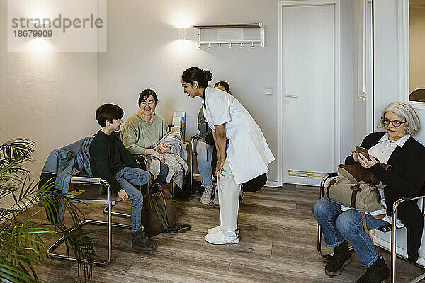 Ärztin mit Händen auf dem Knie im Gespräch mit einem Jungen  der mit seiner Mutter im Wartezimmer sitzt