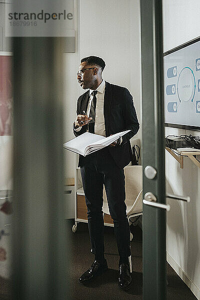 Reifer männlicher Unternehmer  der ein Notebook während eines Geschäftstreffens im Büro hält  gesehen durch die Türöffnung
