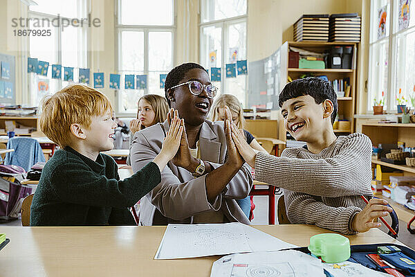 Glückliche Lehrerin und Jungen geben sich High-Fives  während sie am Schreibtisch im Klassenzimmer sitzen