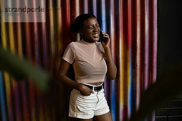 Lächelnde junge afroamerikanische Frau im Gespräch mit Handy auf Wand aus Metall Farben Hintergrund