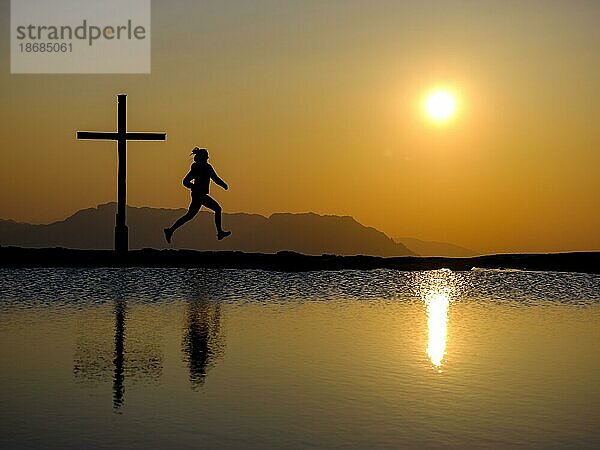 Silhouette einer Läuferin mit Gipfelkreuz bei Sonnenuntergang  Spiegelung im Wasser  Trattberg  Bad Vigaun  Land Salzburg  Österreich  Europa