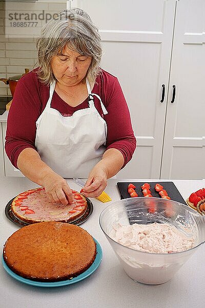Eine weißhaarige Konditorin mit weißer Schürze bereitet in ihrer Küche eine Erdbeer Torte zu