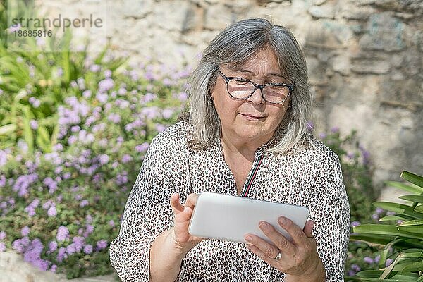 Ältere weißhaarige Frau mit Brille  die in einem Park mit Blumen sitzt und ihr digitales Tablet konsultiert
