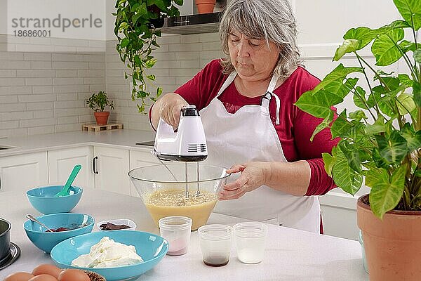 Köchin in weißer Schürze schlägt Eier in einer Schüssel mit einem elektrischen Mixer auf  um einen Erdbeerkuchen herzustellen