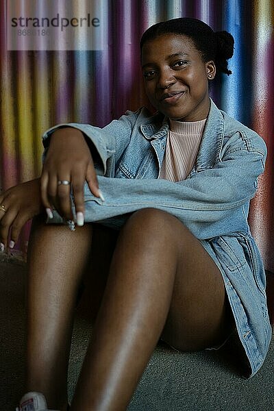 Hübsches junges afroamerikanisches Mädchen  das lächelnd in die Kamera blickt und mit dem Rücken an eine bunte Metallwand gelehnt sitzt