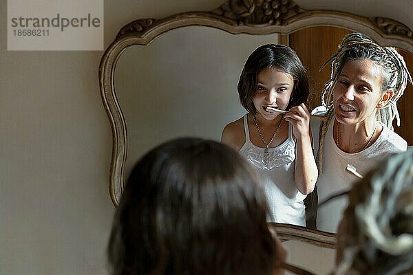 Mädchen schaut in den Spiegel und putzt sich die Zähne  während ihre Mutter sie anlächelt. Bild reflektiert im Badezimmerspiegel