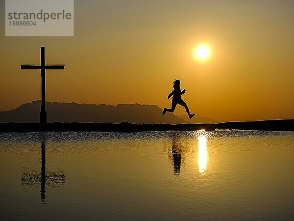 Silhouette einer Frau mit Freudensprung neben Gipfelkreuz bei Sonnenuntergang  Spiegelung im Wasser  Trattberg  Bad Vigaun  Land Salzburg  Österreich  Europa