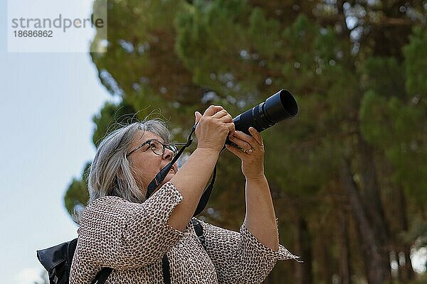 Ältere Frau mit weißen Haaren und Brille  die ein Foto macht und durch den Sucher ihrer Kamera schaut  mit Bäumen im Hintergrund