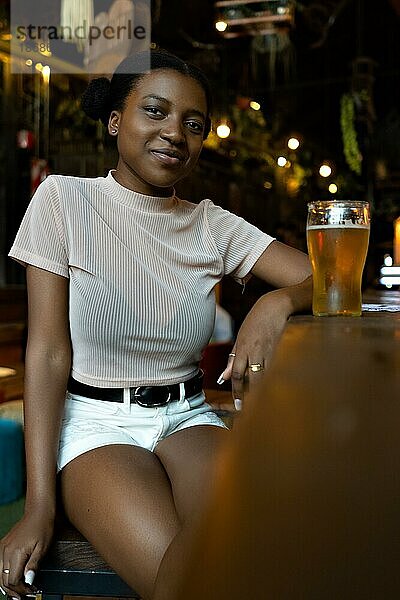 Vertikales Porträt einer jungen schwarzen Frau mit Afrofrisur  die in einer Bar sitzt und in die Kamera schaut