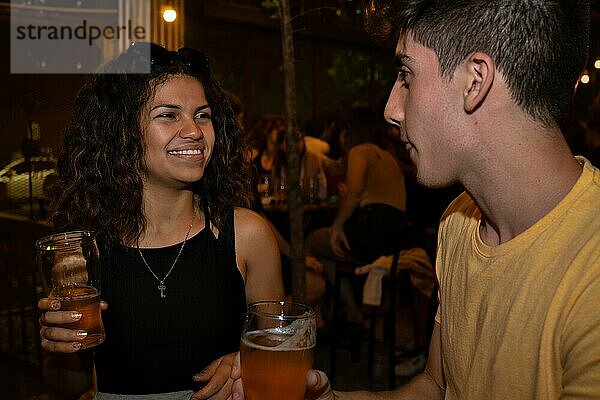 Junge lateinamerikanische Frau lächelt und plaudert mit einem jungen Mann in einer Bar und trinkt Bier. Treffen bei einem Online Date. Unscharfer Hintergrund