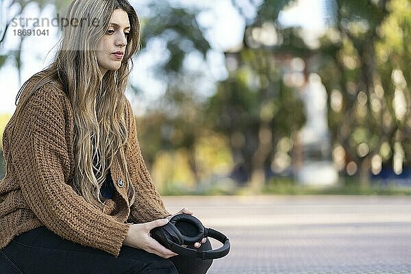 Junge Frau sitzt mit besorgtem Gesichtsausdruck und verlorenem Blick in einem Park und hält Kopfhörer in der Hand