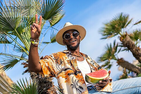 Schwarzer ethnischer Mann  der den Sommerurlaub am Strand genießt  einen Hut und eine Wassermelone trägt und das Okay gibt