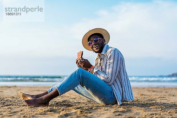 Porträt eines schwarzen ethnischen Mannes genießen Sommerurlaub am Strand essen eine Wassermelone  Sitting on the sand lächelnd