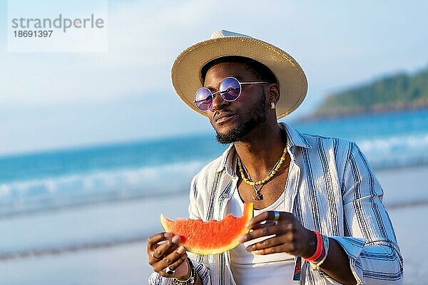 Porträt eines schwarzen ethnischen Mannes  der den Sommerurlaub am Strand genießt und eine Wassermelone isst und einen Sonnenschirm und eine Sonnenbrille trägt