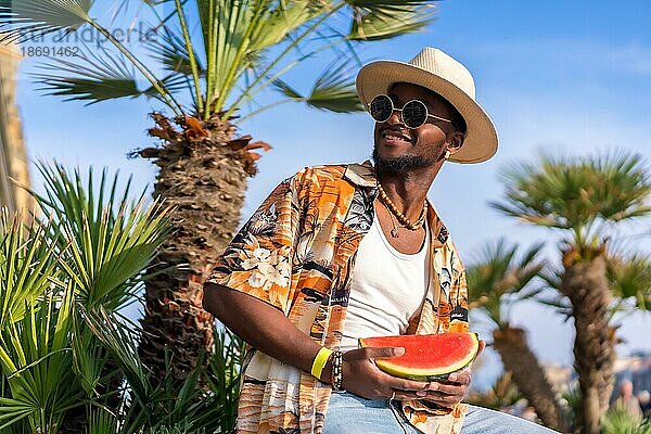 Schwarzer ethnischer Mann genießt Sommerurlaub am Strand  trägt einen Hut und eine Wassermelone