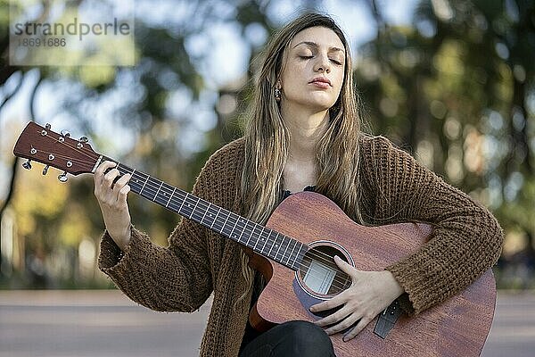 Verträumte und berührte zarte blonde Frau  schließt die Augen und spielt Gitarre mit zufriedenem freundlichem Lächeln in einem Park