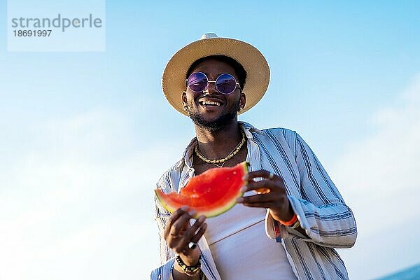 Schwarzer ethnischer Mann genießt Sommerurlaub am Strand und isst eine Wassermelone
