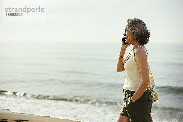 Lächelnde Frau mit der Hand in der Tasche  die am Strand mit dem Smartphone spricht