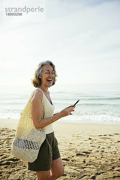 Glückliche Frau mit Netztasche und Smartphone am Strand