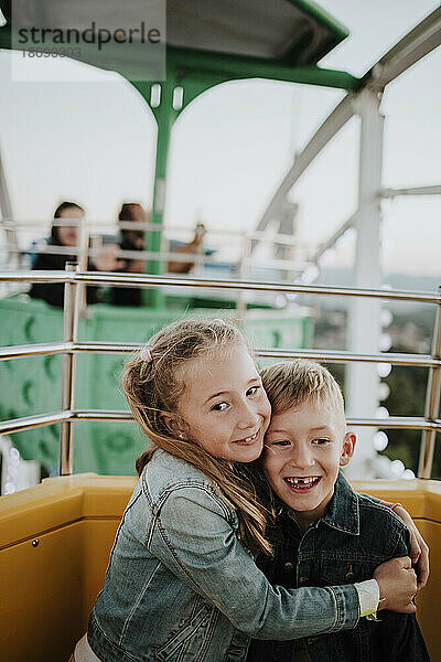 Lächelndes Mädchen umarmt Bruder  der im Riesenrad im Vergnügungspark sitzt