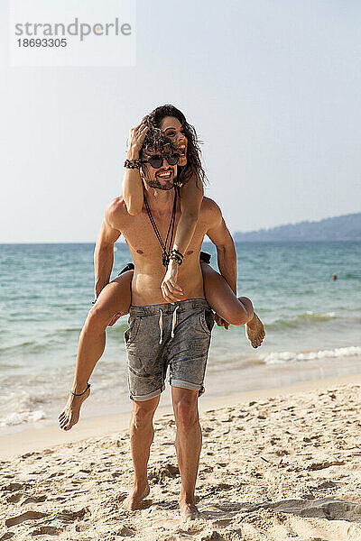 Glücklicher Mann  der seiner Freundin Huckepackfahrt am Strand gibt