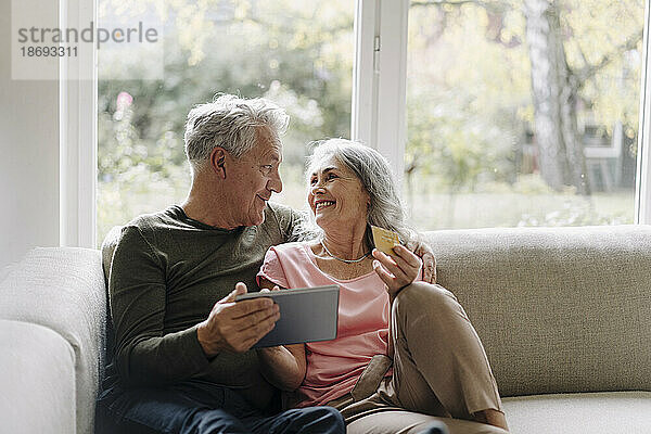 Happy senior couple relaxing on couch at home using tablet for online shopping