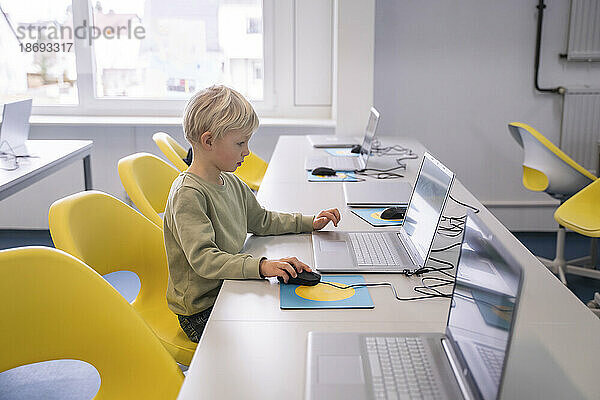 Blonder Junge sitzt mit Laptop auf Stuhl in der Schule